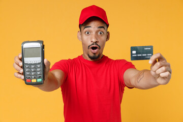 Delivery employee african man 20s in red cap t-shirt uniform work courier service hold wireless bank payment terminal to process and acquire credit card payments isolated on yellow background studio.