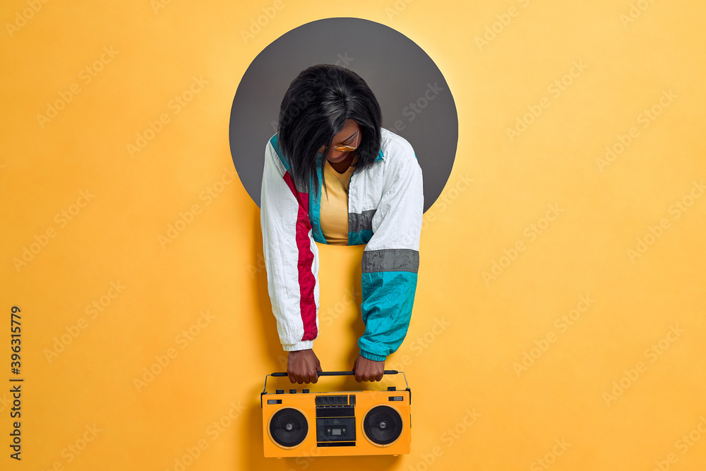 Wall mural Young  woman in retro jacket holding boombox in a circle hole in a bright wall. Copy space.