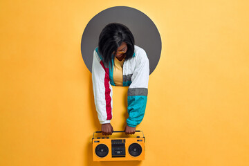 Young  woman in retro jacket holding boombox in a circle hole in a bright wall. Copy space.