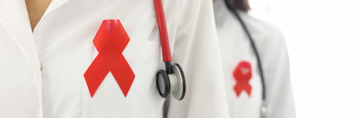 Doctors stand with phonendoscope around their neck and red ribbon on uniforms close-up. World aids day concept.