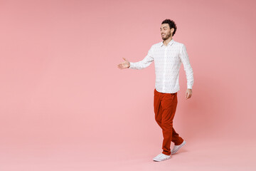 Fototapeta na wymiar Full length side view of excited young bearded man 20s wearing basic casual white shirt standing with outstretched hand for greeting isolated on pastel pink color wall background studio portrait.