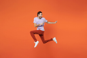 Fototapeta na wymiar Full length side view of excited screaming young bearded man 20s wearing casual violet t-shirt jumping pointing index fingers aside isolated on bright orange color wall background studio portrait.