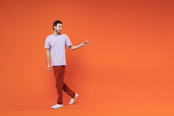 Fototapeta na wymiar Full length side view of amazed young bearded man 20s in casual violet t-shirt standing pointing index finger aside on mock up copy space isolated on bright orange color background studio portrait.
