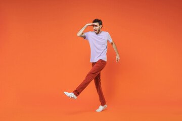 Full length of excited cheerful young bearded man 20s in casual violet t-shirt standing holding hand at forehead looking far away distance isolated on bright orange color background studio portrait.