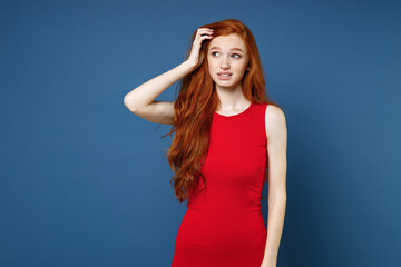 Preoccupied puzzled pensive confused young redhead woman 20s wearing bright red elegant evening dress standing put hand on head looking aside isolated on blue color wall background studio portrait.