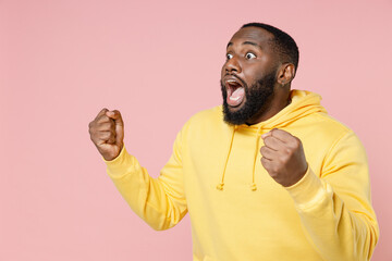 Shocked young african american man 20s wearing basic yellow streetwear hoodie standing doing winner gesture clenching fists looking aside isolated on pastel pink color wall background studio portrait.