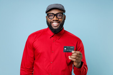 Smiling attractive handsome young bearded african american man 20s wearing casual red shirt eyeglasses cap standing hold credit bank card isolated on pastel blue color wall background studio portrait.