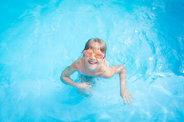 Fototapeta na wymiar a little girl in a bathing suit and pink swimming glasses swims in a pool with blue water
