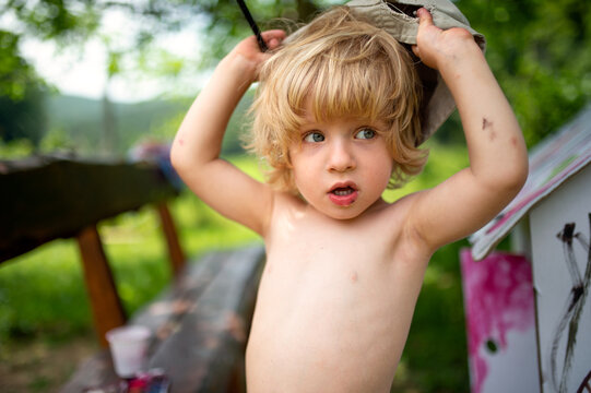 Topless small blond boy with hat painting outdoors in summer.