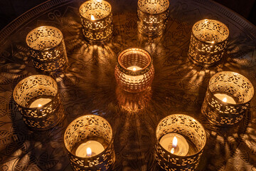 Nine lighted decorative candles, 8 small metal candlesticks surrounding a glass candlestick in the middle of a round metal table, the lighting reflects off the table on a magical December night
