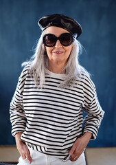 Portrait of senior woman with sunglasses and beret standing indoors against dark background.