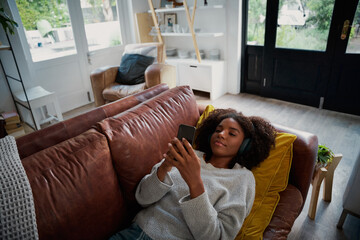 Lazy young woman lying down on couch wearing headphones looking at smart phone screen listening music online