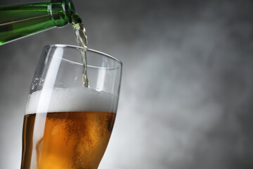 Pouring cold tasty beer from bottle into glass on grey background, closeup. Space for text