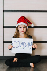 An angry and frowning preschool girl, a child in a red Santa Claus hat, sits and holds a piece of paper with the inscription covid -19. Quarantine on children's holidays at Christmas and New Year.