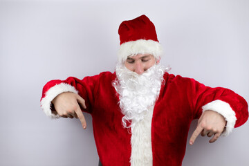Man dressed as Santa Claus standing over isolated white background surprised, looking down and pointing down with fingers and raised arms