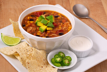 Bowl of chicken taco soup, served with tortilla chips, chili peppers, and sour cream in a white bowl and plate