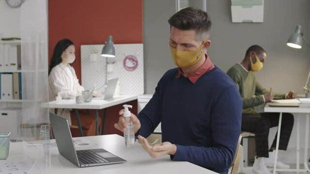 Waist up of middle-aged Caucasian male person in yellow cloth mask sitting by desk in coworking space, using hand sanitizer, typing on laptop keyboard. Busy colleagues on background doing their job
