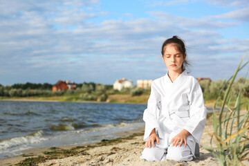 Naklejka na ściany i meble Cute little girl in kimono meditating near river. Karate practicing