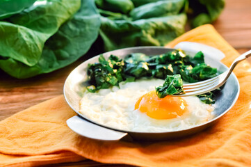 Green cooked spinach with fried egg on iron pan plate. Fried eggs healthy breakfast eating on wooden table background served by orange napkin, fork and spinach leaves, ingredients. Close up