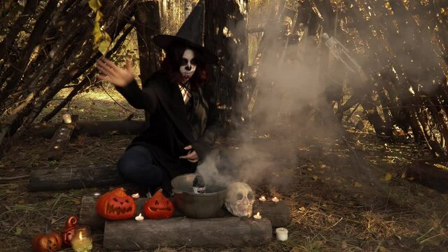 Witch in mantle with santa muerte makeup moves her hand over smoke. Woman is sitting in forest among pumpkins, skull and kettle. Theme of halloween and witchcraft in autumn forest.
