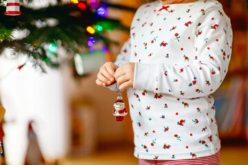 Adorable toddler girl in pajamas decorating Christmas tree with toy in cute hands. Little child in nightwear standing by Xmas tree. celebration of traditional family winter holiday