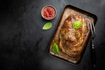 Baked pork meat in a baking pan on dark background, top view
