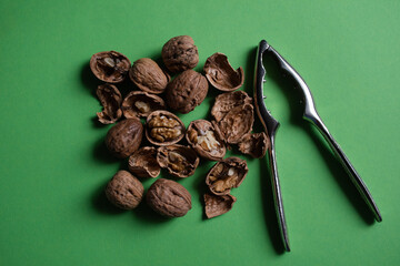Cracked and whole walnuts and shells next to a silver nutcracker on a plain green background