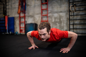 Determined fit young Hispanic man in red short-sleeved shirt doing push-ups in modern indoor gym. Healthy and active sport lifestyle