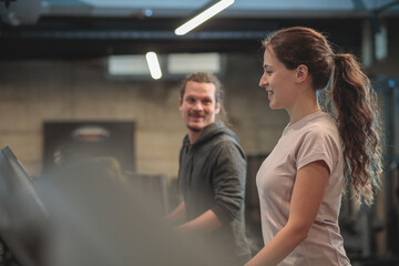 Smiling and happy young mixed ethnicity couple jogging on treadmills in modern indoor gym and talking with each-other