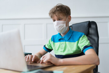 A Caucasian boy in a disposable face mask during his quarantine online classes. He's typing the text and listening to the teacher
