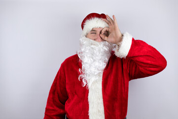 Man dressed as Santa Claus standing over isolated white background doing ok gesture shocked with smiling face, eye looking through fingers