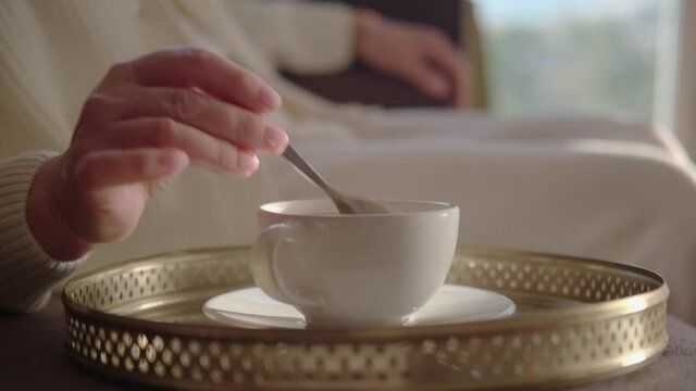 Hand of a pensioner is slowly stirring sugar in a white cup of coffee