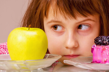 Close uup young cute beautiful girl having hard choice between healthy and unhealthy food. She choosing between apple fruit and tasty cake. Horizontal image.