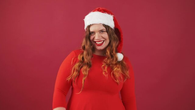 Smiling plus size lady in Christmas red hat, waving hands in hello gesture, greeting friends, red studio background.