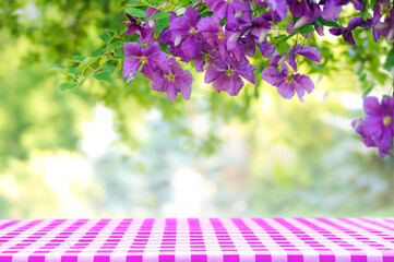 Natural summertime cooking template - fresh green leaves and violet flowers of clematis over an empty empty table with  violet checkered tablecloth on a sunny day with copy space.