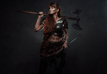 Barbaric and grimy woman dressed in dark armour holding two axes and posing in dark background.