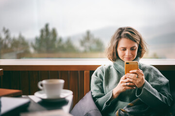 Young woman in a green sweater using a mobile phone near window in country house with a view of the mountains and pine trees - Powered by Adobe