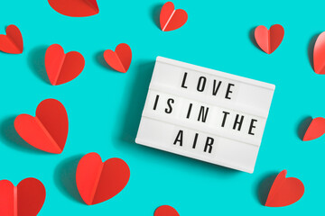 Festive background with glowing lightbox with text Love is in the Air and red paper hearts on bright blue background for Valentine's Day. Flatlay. Top view.