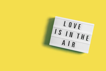 Festive background with glowing lightbox with text Love is in the Air on yellow illuminating background for Valentine's Day. Flatlay for 14th February. Top view. Copyspace.