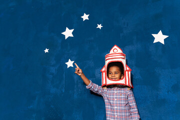African american kid boy with rocket helmet on head on blue studio background with white stars....