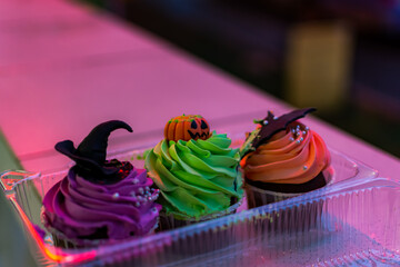 Colorful cakes for Halloween. Set of birthday cakes in a plastic box in neon lighting. Beautiful pumpkin, witch and bat cake. Soft evening lighting. Hello, Halloween. Copy space