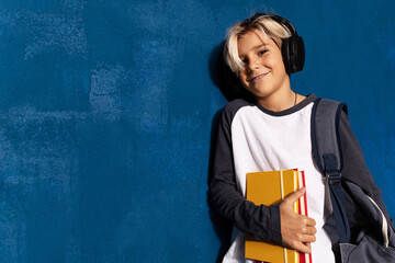 Smiling schoolboy in wireless headphones with textbooks and backpack posing on blue background with blank space for advertising. Stylish teen boy in headset listening lesson, music or podcast.