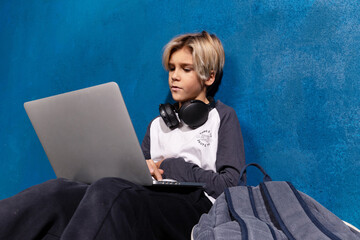 Schoolboy with laptop and wireless headphones doing homework sitting on floor with backpack. Focused stylish teen boy learning lessons using internet. E-learning concept.