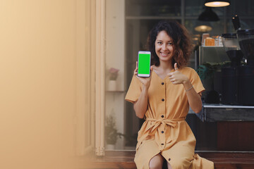 Fototapeta premium Mockup image of a woman wearing yellow dress holding and showing mobile phone with blank green screen at coffee shop