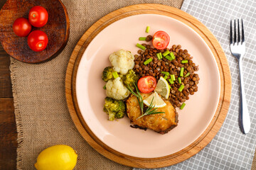 Baked fish with broccoli and lentils in a beige plate on a round stand next to tomatoes and lemon.