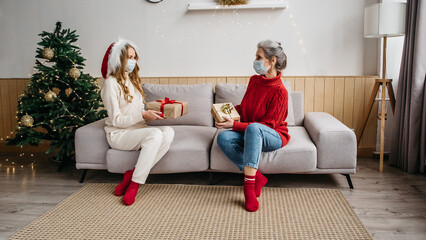 Health protection while celebrating Christmas and New Year 2021 concept. Senior woman and her daughter wearing face masks, taking distance and sitting on the couch in Christmas decorated room