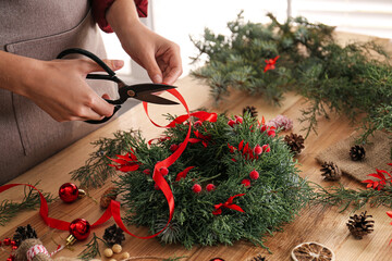 Florist making beautiful Christmas wreath with berries and red ribbon at wooden table indoors, closeup