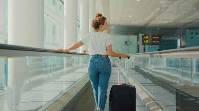Rear view on beautiful authentic young female, woman traveller with simple black suitcase stand on moving walkway or travelator in empty airport terminal. Travel in times of covid restrictions