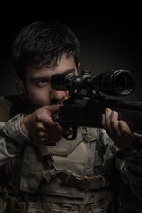 young man with a beard standing, holding and aiming a rifle, uses an earphone and camouflage...