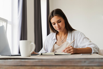Attractive business woman sits at table in laptop, smartphone lies on table, makes notes with a pencil in notebook.